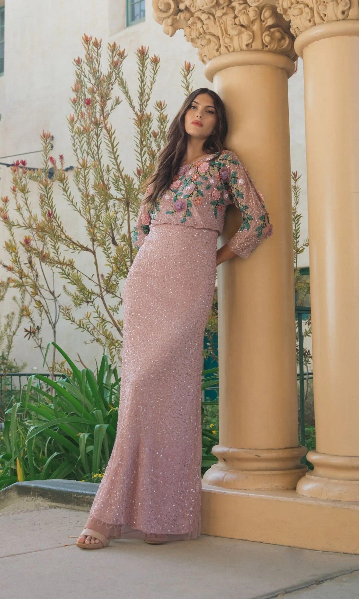 Wearing the Mac Duggal 9341 long formal dress, a woman stands leaning against an ornate column outdoors, surrounded by lush green plants. The gown features light pink sequins and intricate floral embroidery.